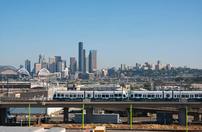 Link light rail trains traveling on an elevated guideway on a clear day, with the Seattle city skyline in the background.