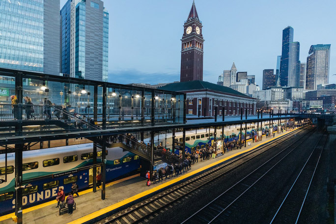 Passengers standing on the platform, waiting to board the Sounder Train in downtown Seattle at dusk. The Seattle skyline, including the Union Station clock tower, is illuminated in the distance.